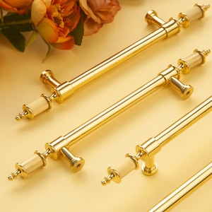 Op de afbeelding: Drie gouden deurgrepen met een decoratieve, geribbelde, crèmekleurige knop aan elk uiteinde. De grepen bevinden zich op een lichtgele ondergrond.