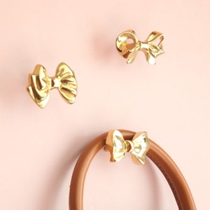 Pode incluir: Três ganchos dourados em forma de laço, dois sobre fundo rosa e um sobre uma tira de couro marrom.