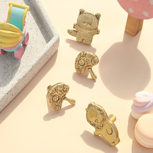 May include: A collection of gold-colored cabinet knobs in various shapes, including a bear, mushroom, and lion. The knobs are arranged on a light-colored surface, with a toy stroller and a mushroom-shaped decoration in the background.