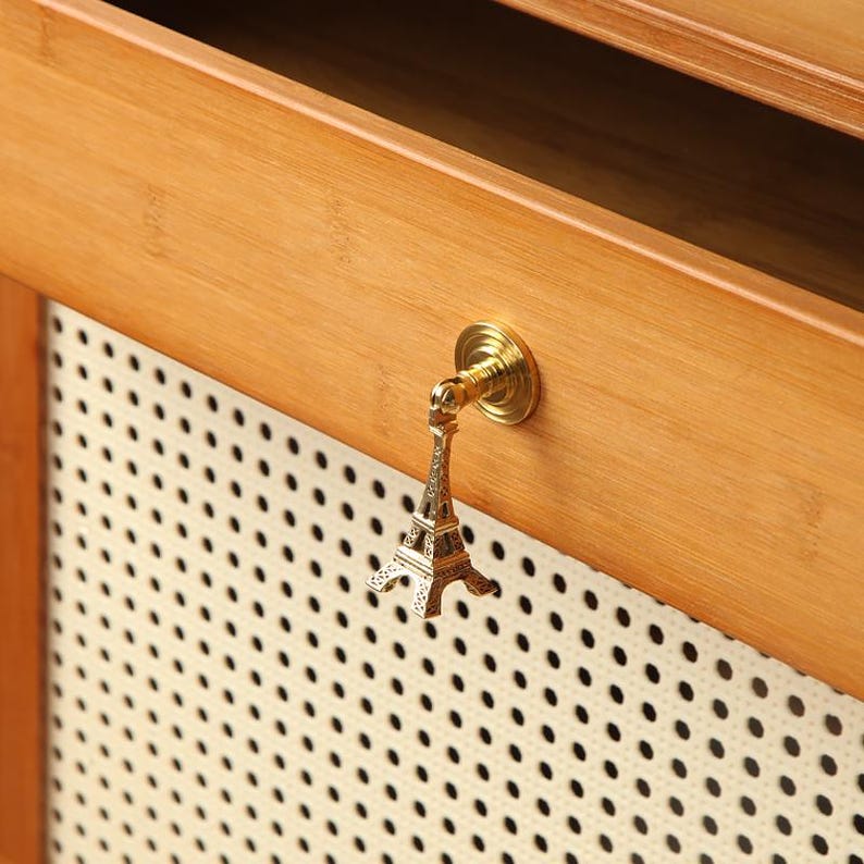 Peut inclure: Un bouton de meuble dor&eacute; en forme de Tour Eiffel fix&eacute; sur une surface en bois brun clair. Le bouton est mont&eacute; sur une base ronde dor&eacute;e. L'arri&egrave;re-plan pr&eacute;sente un panneau perfor&eacute; blanc.