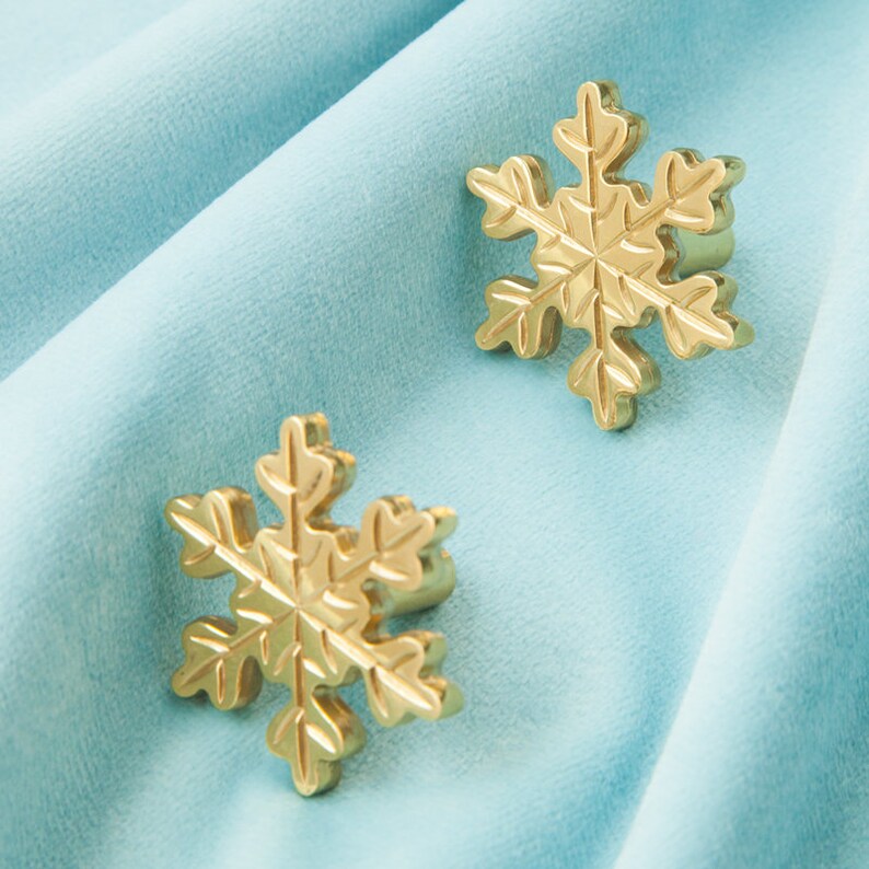 Op de afbeelding: Twee gouden sneeuwvlok-vormige ornamenten op een lichtblauwe fluwelen achtergrond.