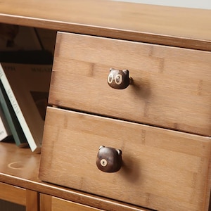 May include: A light brown wooden drawer unit featuring two horizontal drawers. Each drawer is equipped with a dark brown wooden knob shaped like an animal head. The top knob depicts a cat face, while the bottom knob depicts a bear face.