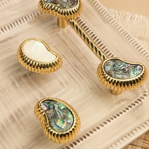 May include: Three gold cabinet knobs with a decorative rope design. The knobs are made of metal and have a mother-of-pearl inlay. The knobs are in a variety of shapes, including a teardrop, a round shape, and a rectangular shape.