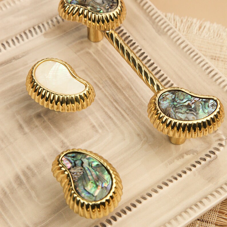 May include: Three gold cabinet knobs with a decorative rope design. The knobs are made of metal and have a mother-of-pearl inlay. The knobs are in a variety of shapes, including a teardrop, a round shape, and a rectangular shape.