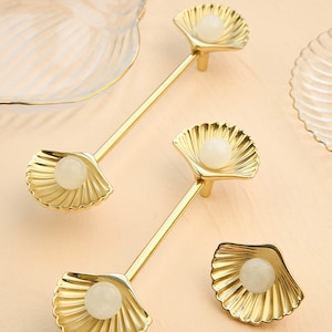 May include: Two gold cabinet knobs with a seashell design. Each knob features a white pearl-like centre and a detailed shell pattern.