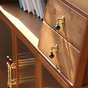 Op de afbeelding: Houten kast met twee lades. Elke lade heeft een goudkleurige handgreep met een donkerbruine bol en een groene kraal. De kast is gemaakt van lichtbruin hout en heeft glazen deuren.