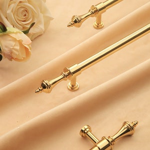 Op de afbeelding: Drie gouden kastdeurgrepen met sierlijke, decoratieve uiteinden. De grepen bevinden zich op een lichtbeige stoffen achtergrond.