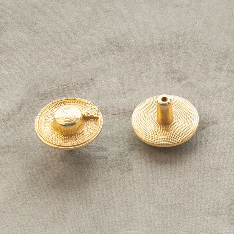 May include: Two gold-toned cabinet knobs. One knob is shaped like a hat with a small flower detail. The other knob is round with a raised centre.