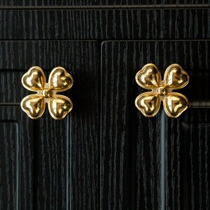 May include: Two gold-colored four-leaf clover shaped cabinet knobs on a black wooden surface.