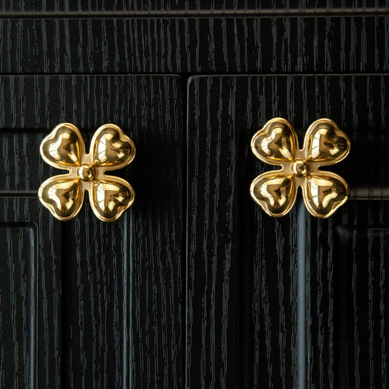 May include: Two gold-colored four-leaf clover shaped cabinet knobs on a black wooden surface.