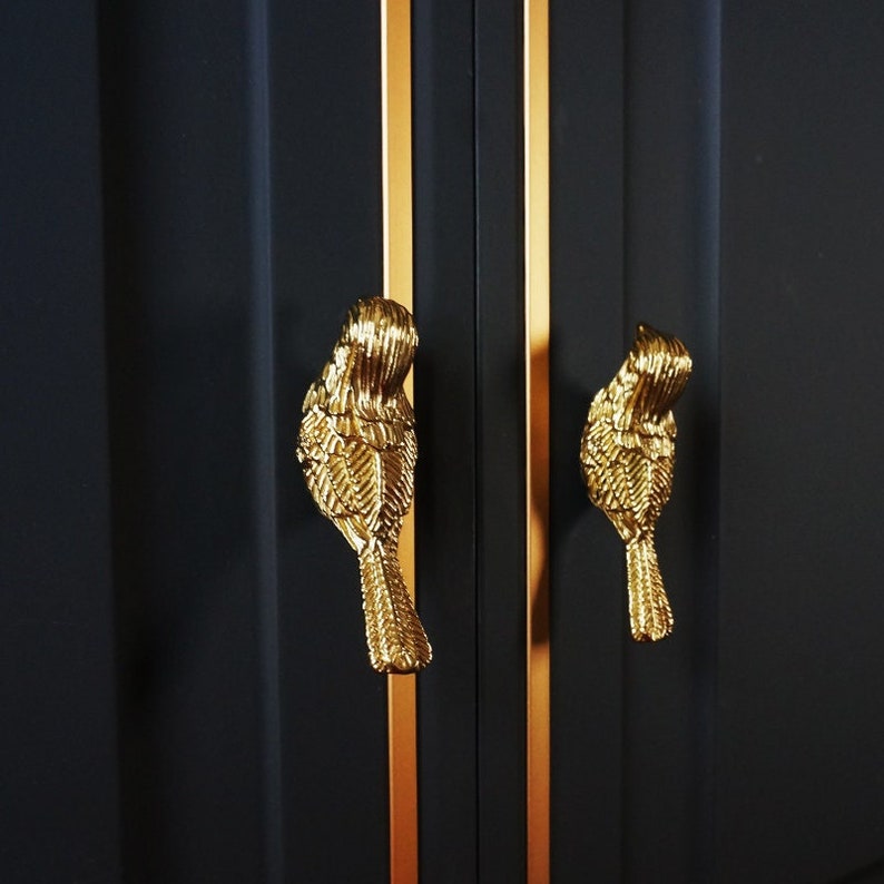 May include: Two gold bird-shaped cabinet knobs with detailed feather designs against a black background with gold trim.