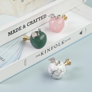 K&ouml;nnte beinhalten: Drei dekorative, apfelf&ouml;rmige Schrankkn&ouml;pfe in Gr&uuml;n, Rosa und wei&szlig;em Marmor. Jeder Knopf hat einen goldfarbenen Stiel und ein silberfarbenes Blattdesign. Die Kn&ouml;pfe werden auf einem wei&szlig;en Buch mit dem Text "MADE & CRAFTED" und "THE KINFOLK HOME" pr&auml;sentiert.