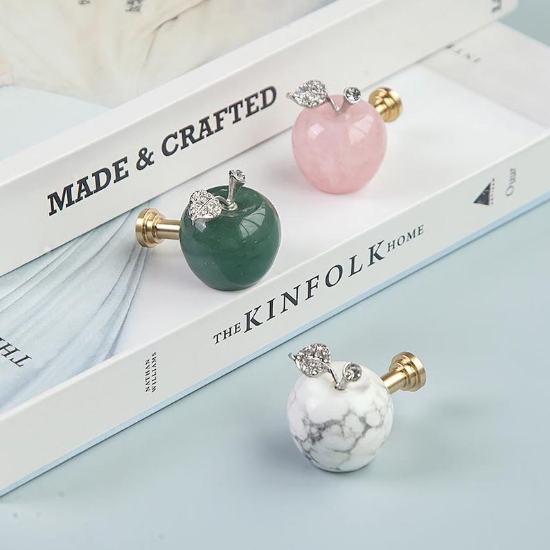 K&ouml;nnte beinhalten: Drei dekorative, apfelf&ouml;rmige Schrankkn&ouml;pfe in Gr&uuml;n, Rosa und wei&szlig;em Marmor. Jeder Knopf hat einen goldfarbenen Stiel und ein silberfarbenes Blattdesign. Die Kn&ouml;pfe werden auf einem wei&szlig;en Buch mit dem Text "MADE & CRAFTED" und "THE KINFOLK HOME" pr&auml;sentiert.