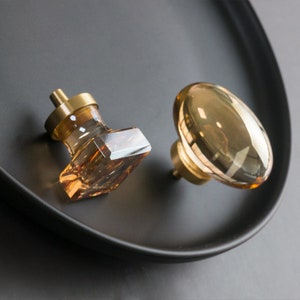 Op de afbeelding: Twee amberkleurige glazen kastknoppen met messing bases. De ene knop is vierkant en de andere rond.