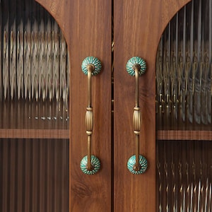 May include: Close-up of a wooden cabinet door with two ornate handles. The handles are brass-colored with a green and white floral design on the ends. The cabinet doors are made of dark wood and have textured glass panels.