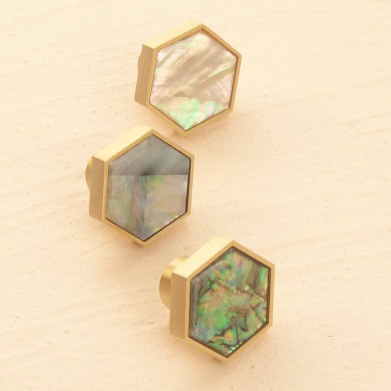 May include: Three hexagonal cabinet knobs with a gold finish and iridescent mother of pearl inlays. The knobs are arranged in a triangular pattern on a light beige surface.