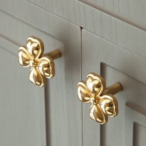 May include: Two gold-toned flower-shaped cabinet knobs with a simple design. The knobs are mounted on a light gray cabinet door.