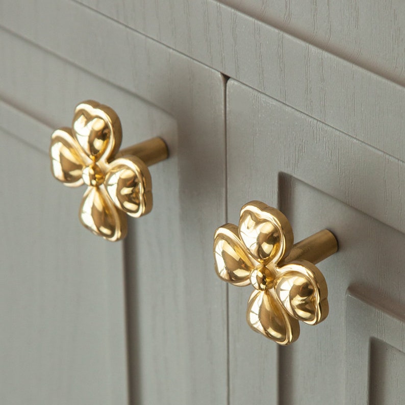 May include: Two gold-toned flower-shaped cabinet knobs with a simple design. The knobs are mounted on a light gray cabinet door.