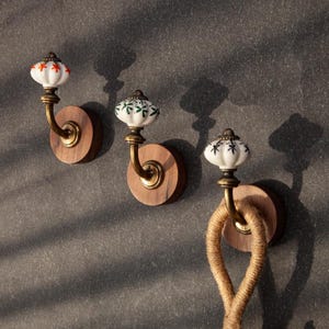 May include: Three decorative wall hooks with ceramic knobs and brass fittings. The knobs have floral patterns in red, green, and black on a white base. Each hook is fixed to a round wooden base, with a rope hanging from one.