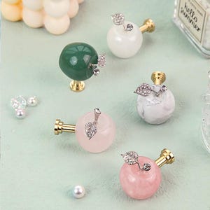 K&ouml;nnte beinhalten: Dekorative M&ouml;belkn&ouml;pfe in Apfelform in verschiedenen Farben: Gr&uuml;n, Wei&szlig;, Marmor und Rosa. Jeder Knopf hat einen goldfarbenen Stiel und ein dekoratives Blatt mit Kristallakzenten. Geeignet f&uuml;r M&ouml;bel und Heimdekoration.