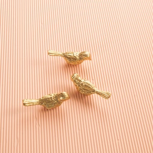 May include: Three gold-colored bird figurines on a pink textured background. The birds are facing different directions and have detailed feather patterns.