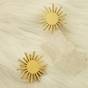 May include: Two gold sunburst-shaped ornaments with a textured surface, sitting on a white fluffy surface with two clear quartz crystals.