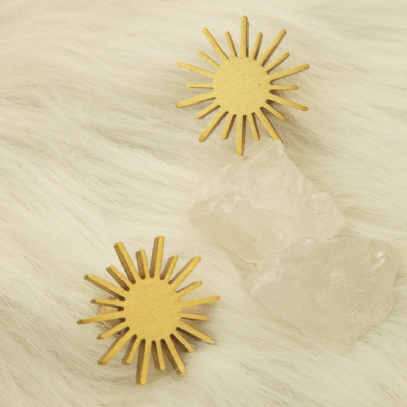 May include: Two gold sunburst-shaped ornaments with a textured surface, sitting on a white fluffy surface with two clear quartz crystals.