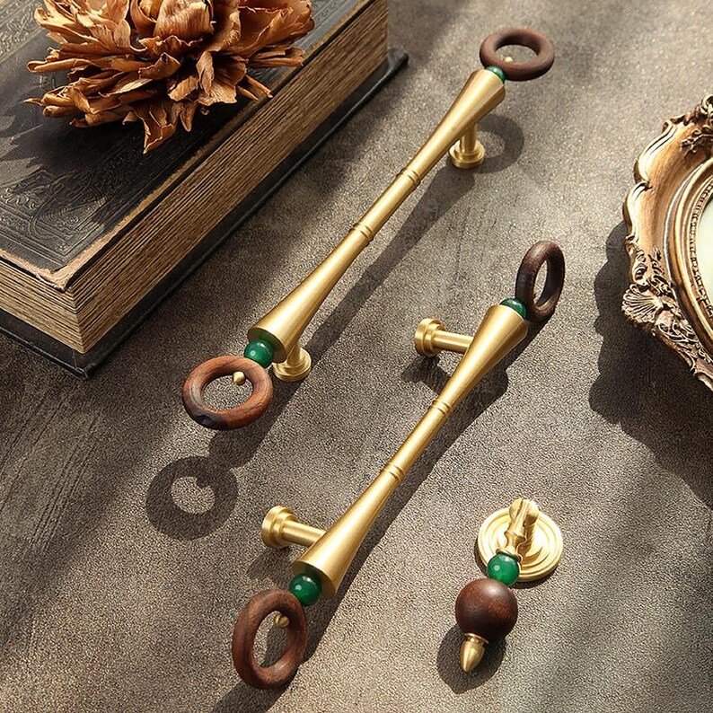 Peut inclure: Un ensemble de poign&eacute;es de meubles en laiton et un bouton. Les poign&eacute;es ont un design long et cylindrique avec une poign&eacute;e en anneau de bois et un accent de perle verte. Le bouton a un design similaire. La quincaillerie est pr&eacute;sent&eacute;e sur une surface neutre.