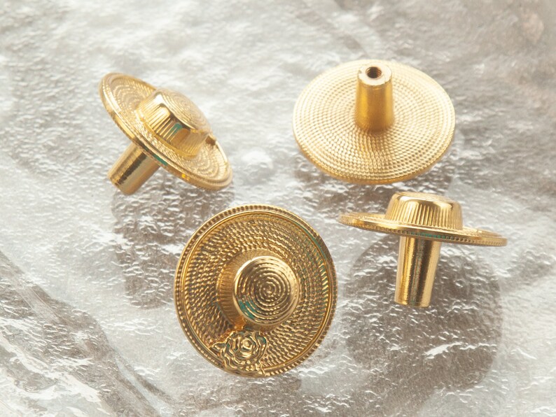 May include: Four gold-toned cabinet knobs with a decorative, textured, circular design resembling a hat. The knobs have a cylindrical stem.