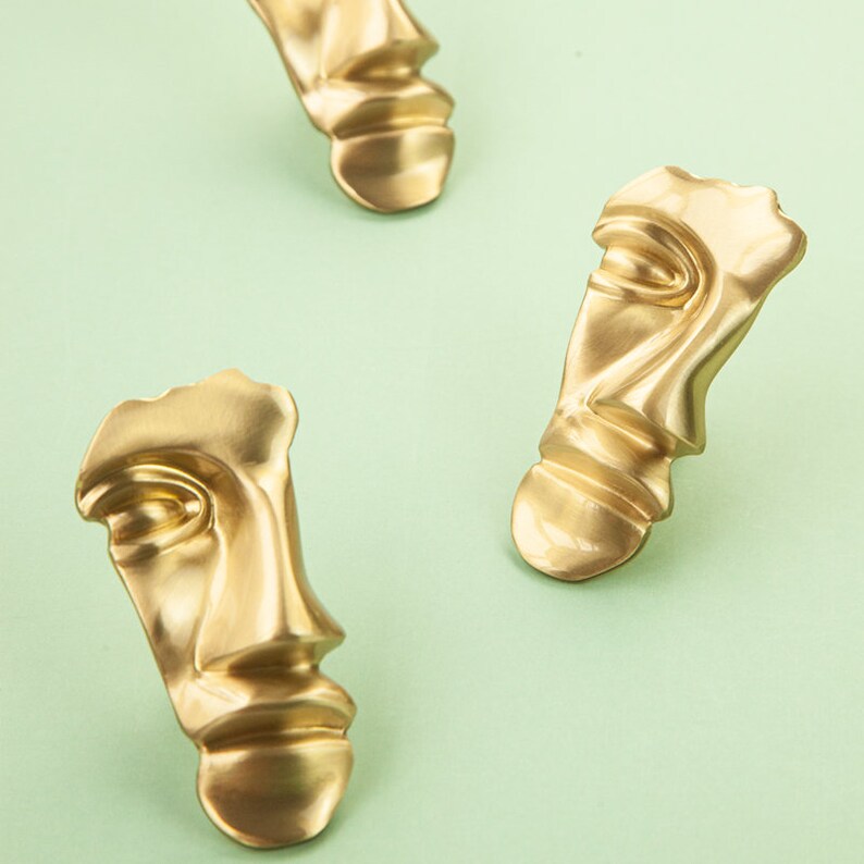 May include: Three gold-colored abstract face sculptures with a textured surface. The sculptures are arranged in a row on a light green background.