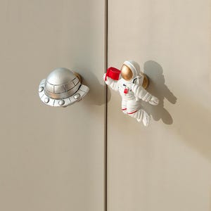 Könnte beinhalten: Zwei Schrankknöpfe: eine silberne UFO und ein Astronaut in einem weißen Raumanzug mit goldenen Akzenten und einer roten Box. Die Knöpfe sind an einer hellbeigen Schranktür angebracht und bilden ein dekoratives Element zum Thema Weltraum.