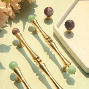 Può includere: Tre tiretti per cassetti dorati con pomelli in pietra preziosa verde e viola. I tiretti hanno un design decorativo e scanalato.