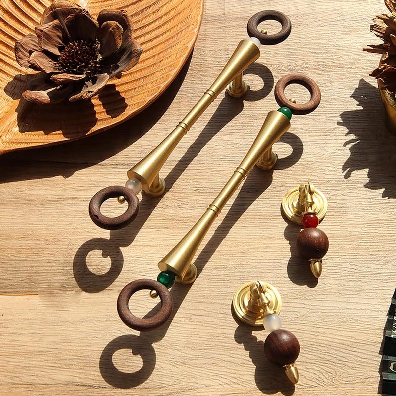 Peut inclure: Tirants et boutons de meuble dor&eacute;s avec des accents en bois. Les tirants ont un design long et effil&eacute; avec une poign&eacute;e en bois en forme d'anneau. Les boutons comportent une perle en bois ronde et une perle rouge plus petite. Quincaillerie d&eacute;corative.