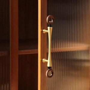 Peut inclure: Gros plan sur une porte de placard en bois avec une poign&eacute;e en laiton. La poign&eacute;e a un design cylindrique avec un anneau en bois brun fonc&eacute; en haut et en bas. La porte de l'armoire est en bois brun clair et poss&egrave;de un panneau de verre nervur&eacute;.