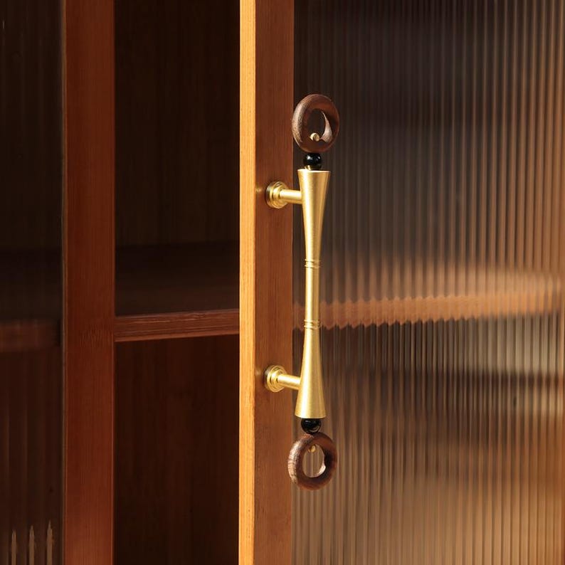 Peut inclure: Gros plan sur une porte de placard en bois avec une poign&eacute;e en laiton. La poign&eacute;e a un design cylindrique avec un anneau en bois brun fonc&eacute; en haut et en bas. La porte de l'armoire est en bois brun clair et poss&egrave;de un panneau de verre nervur&eacute;.