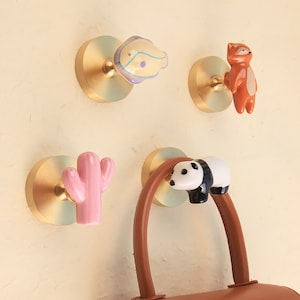 Pode incluir: Quatro ganchos de parede dourados com diferentes designs de cerâmica com temas de animais e natureza. Os ganchos estão montados em uma parede bege claro. Uma bolsa de couro marrom está pendurada em um dos ganchos.