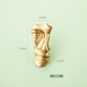 May include: A gold-colored abstract face sculpture with a 36mm height, 74mm width, and 31mm depth. The sculpture weighs 55 grams.