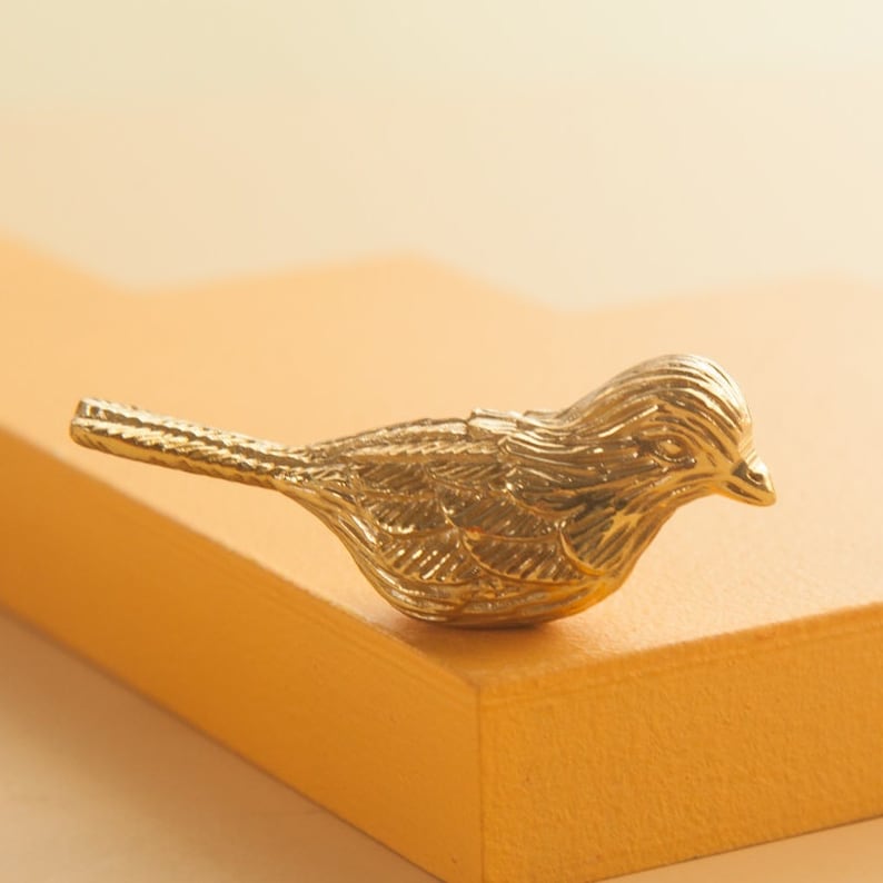 May include: A small, gold-colored bird figurine with detailed feathers. The bird is perched on a light orange surface.