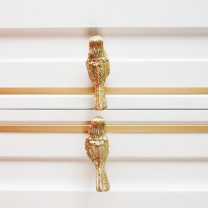 May include: Two gold bird-shaped drawer pulls with detailed feather designs are mounted on a white dresser with gold trim.