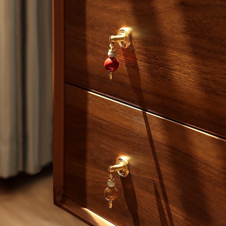 Op de afbeelding: Close-up van een houten kast met twee lades. Elke lade heeft een goudkleurige knop met een rode en witte decoratieve kraal. De kast is gemaakt van donker hout met een zichtbare houtnerf.