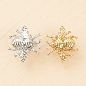 Peut inclure: Deux ornements en m&eacute;tal en forme d'abeille, l'un argent&eacute; et l'autre dor&eacute;, avec des ailes et des corps d&eacute;taill&eacute;s.