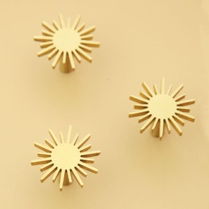 May include: Three gold-toned sunburst-shaped cabinet knobs. Each knob has a circular center and radiating lines.