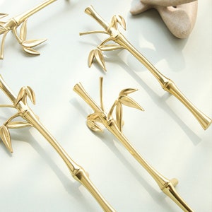 May include: Four gold-toned cabinet pulls shaped like bamboo with a leaf design. The pulls are arranged in a staggered pattern on a white background.