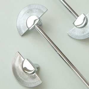 May include: Three silver fan-shaped drawer pulls with a rounded top. The pulls are attached to a silver rod.