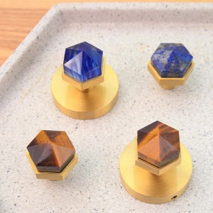 May include: Four hexagonal cabinet knobs with gold bases and different gemstone tops. The gemstones are blue lapis lazuli, tiger's eye, and two different shades of blue.