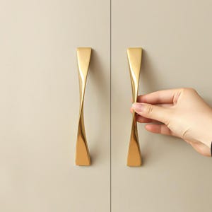 Könnte beinhalten: Zwei goldfarbene Schrankgriffe mit gedrehtem Design. Die Griffe sind an einer hellbeigen Schranktür angebracht. Eine Hand greift nach einem der Griffe. Die Griffe haben eine glatte, reflektierende Oberfläche.
