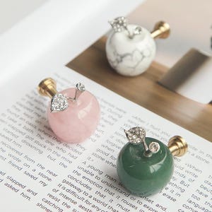 Könnte beinhalten: Drei dekorative Flaschenverschlüsse in Apfelform, rosa, grün und weiß mit grauer Maserung. Jeder hat einen goldfarbenen Stiel und einen Akzent aus Kristallblatt und Edelstein. Die Verschlüsse liegen auf einer Buchseite.