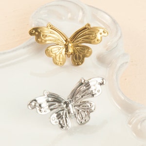 May include: Two butterfly-shaped decorative embellishments. One is gold-coloured, the other is silver-coloured. Both have detailed wing patterns and are displayed on a white, curved surface. The butterflies are likely for crafts or jewellery.