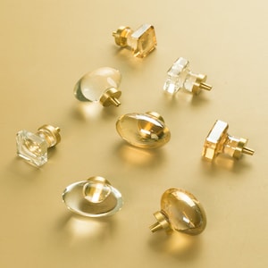 May include: Assortment of seven glass cabinet knobs with gold metal bases. The knobs are in various shapes, including square, oval, and round. Some knobs are clear glass, while others are amber.