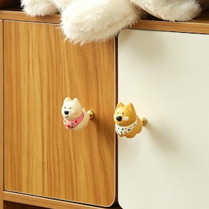 May include: Two decorative cabinet knobs shaped like cartoon dogs. One is cream-colored with a pink and patterned scarf, the other is yellow with a polka-dot scarf. Both knobs have gold-colored stems and are mounted on wooden and white cabinet doors.